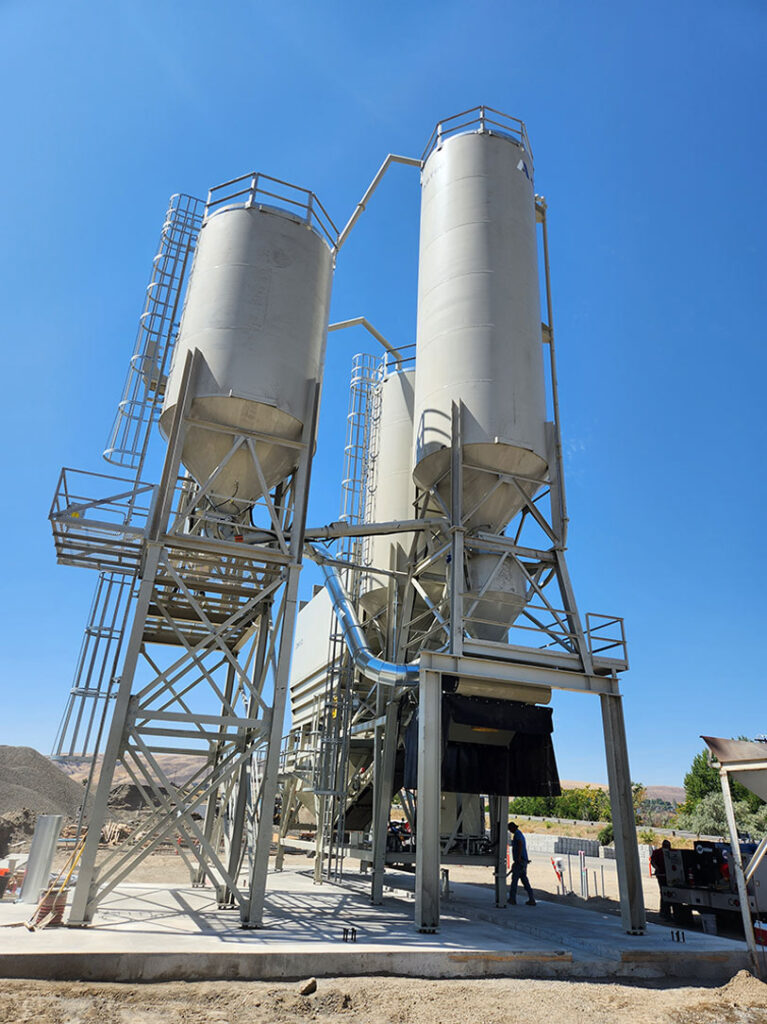 Three large, vertical industrial silos with metal supports stand outdoors on a concrete base under a clear blue sky. A person in safety gear is visible near the base for scale.