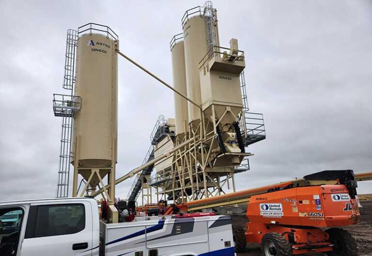 Two large beige industrial silos with connected pipes and platforms stand outdoors under a cloudy sky. In front, a white utility truck and an orange construction lift are parked on dirt ground.