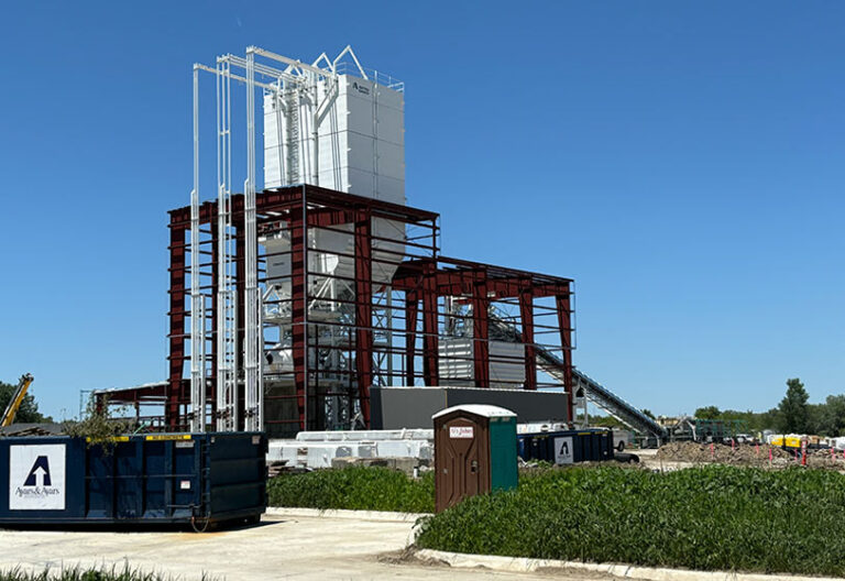 A construction site features a large steel framework with tall white silos and scaffolding. A portable toilet, dumpsters, equipment, and green grass are visible in the foreground under a clear blue sky.