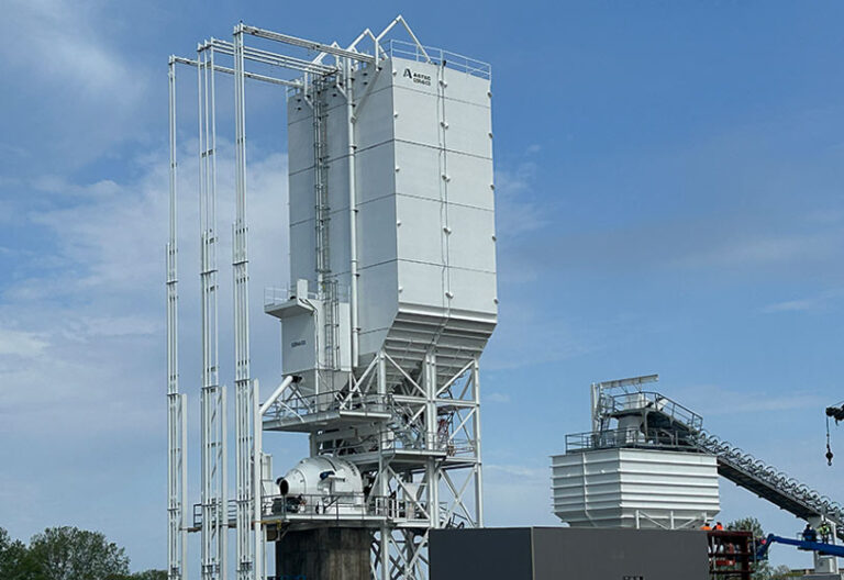 A large industrial structure with tall, white vertical silos and metal framework stands outdoors under a blue sky, with additional machinery and conveyor belts in the background.