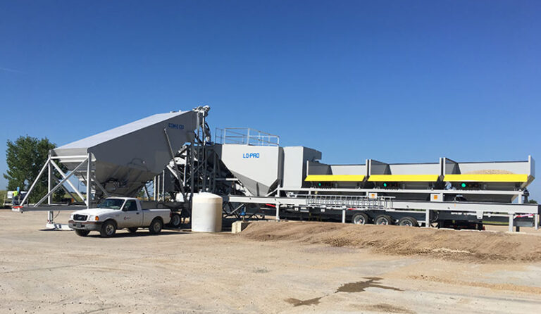 A large portable concrete batch plant with conveyors and storage bins sits on a paved lot under a clear blue sky, with a white pickup truck parked nearby and piles of gravel in the foreground.