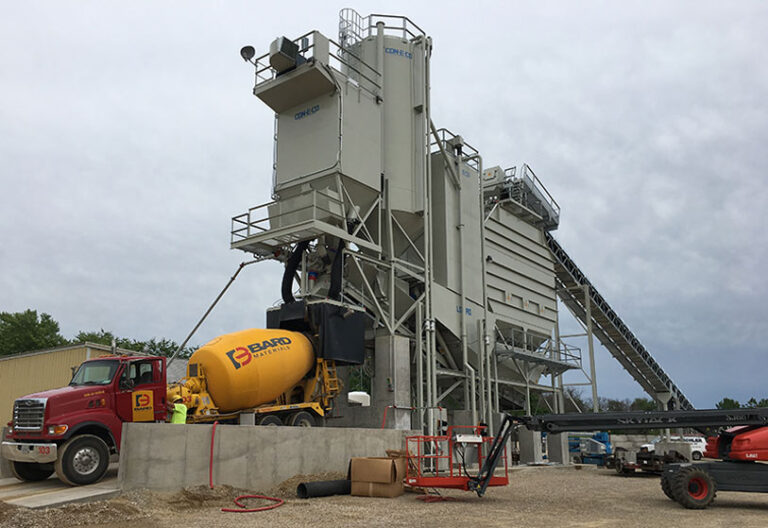 A concrete batching plant with large silos, a conveyor belt, and a yellow BARD mixer truck parked below, next to a red truck and construction workers on a cloudy day.