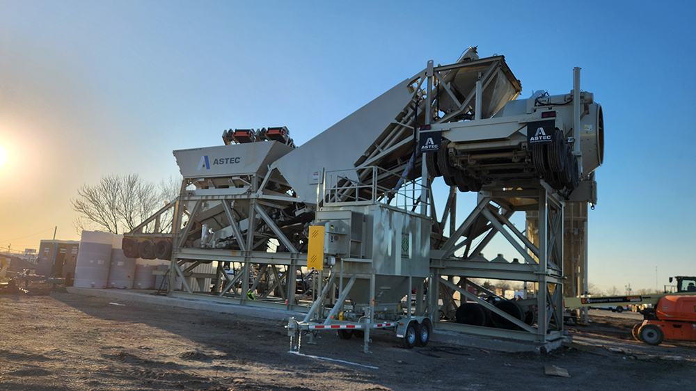 Large industrial asphalt plant machinery with conveyors and storage areas stands outdoors on a construction site at sunrise, with clear skies and bare trees in the background.