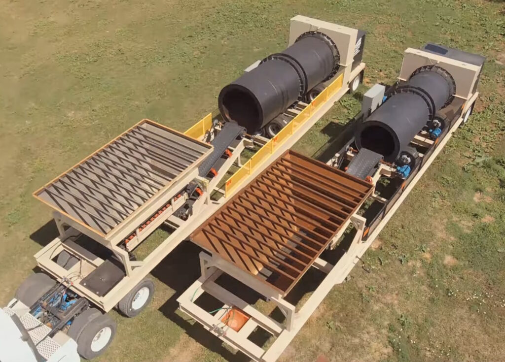 Aerial view of two large mobile trommel screening machines on grassy ground, each with a slanted metal grate and rotating drum, mounted on trailers for transportation.