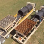 Aerial view of two large mobile trommel screening machines on grassy ground, each with a slanted metal grate and rotating drum, mounted on trailers for transportation.
