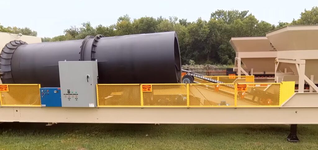 A large industrial sand heater drum machine with a black cylindrical component and yellow safety railings is situated outdoors on grass, with trees visible in the background.