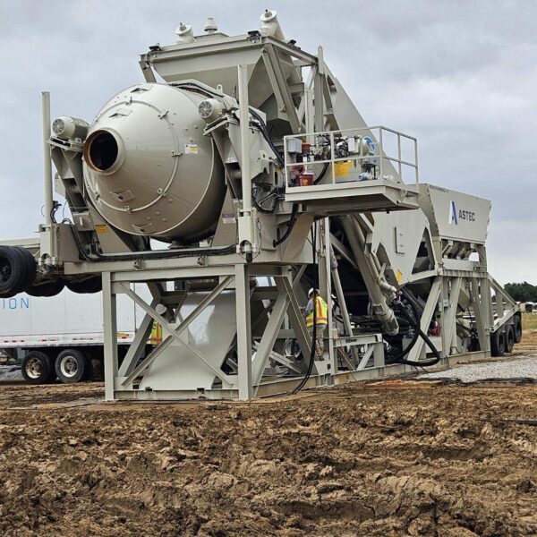 Large industrial asphalt mixing machine with metal framework and conveyor belts, stationed on a muddy construction site under a cloudy sky. Two workers in yellow vests stand nearby for scale.
