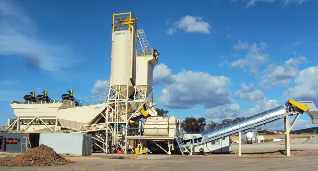 A large, beige industrial concrete batching plant with silos and conveyor belts stands on a wide, dusty lot under a blue sky with scattered clouds. Some construction materials and containers are nearby.