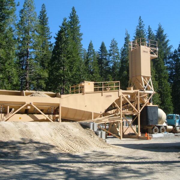 A tan-colored concrete batching plant is set up outdoors near a forest, with piles of sand, processing equipment, and a truck nearby under a clear blue sky.