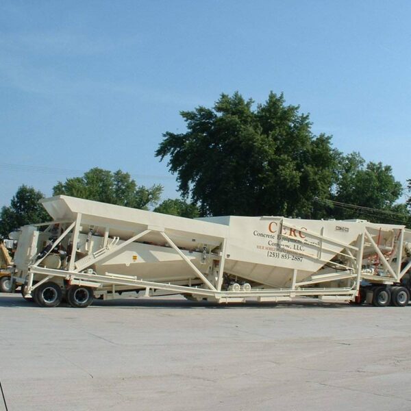 A large portable concrete batch plant mounted on a trailer is parked on a paved lot, surrounded by trees and a blue sky in the background.