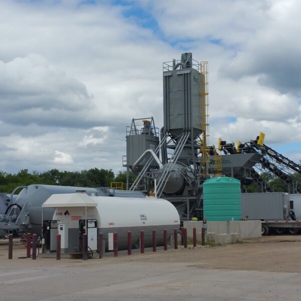 An asphalt mixing plant with industrial equipment, storage tanks, and a fuel station on a cloudy day, set on a gravel lot with trees in the background.