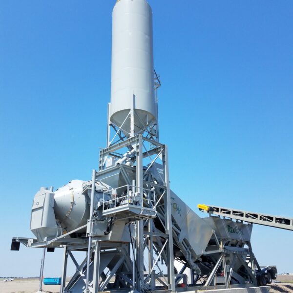 A large concrete batching plant with a tall cylindrical silo and conveyor system stands on a sandy construction site under a clear blue sky.