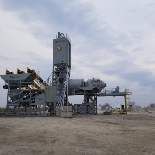 A large industrial concrete batching plant with multiple conveyors and silos stands on a dirt lot under a cloudy sky. Machinery parts are labeled “RexCon.” There are piles of gravel and sparse trees in the background.