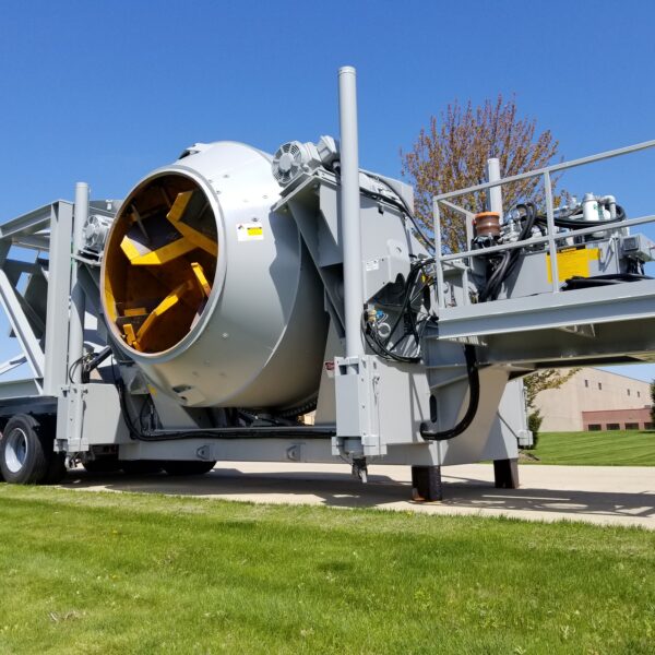 A large, industrial rotary drum machine mounted on a multi-axle trailer sits on grass near a sidewalk under a clear blue sky, with buildings and trees in the background.