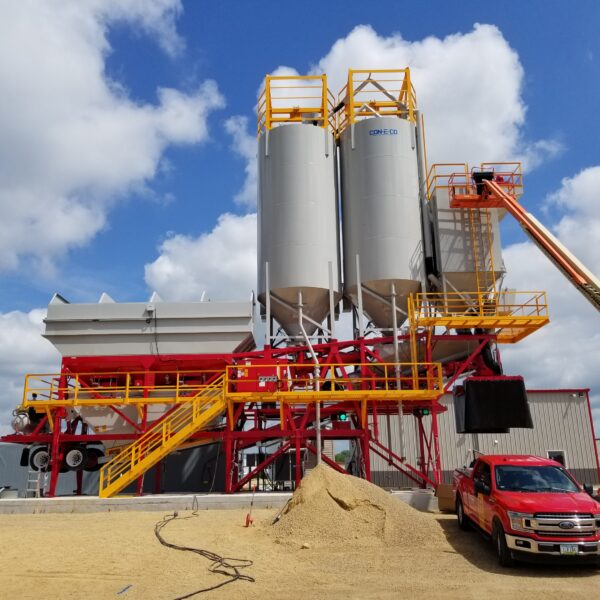 A construction site with silos, yellow and red steel structures, a lifting platform with a worker, a pile of sand, and two red trucks parked in front under a partly cloudy sky.