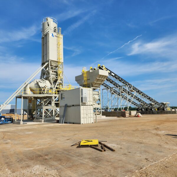 A concrete batching plant with silos, conveyor belts, and storage containers stands on a large, clear construction site under a blue sky with some clouds.