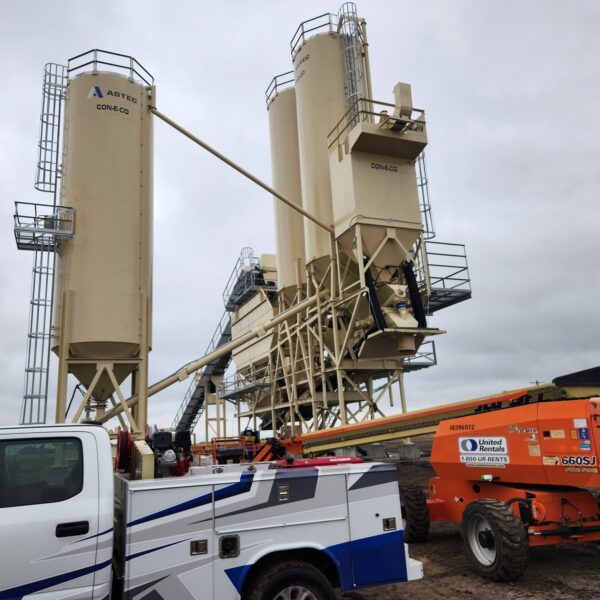 Large industrial silos and machinery stand on a construction site under a cloudy sky. A white utility truck and an orange telescopic lift are parked in the foreground.