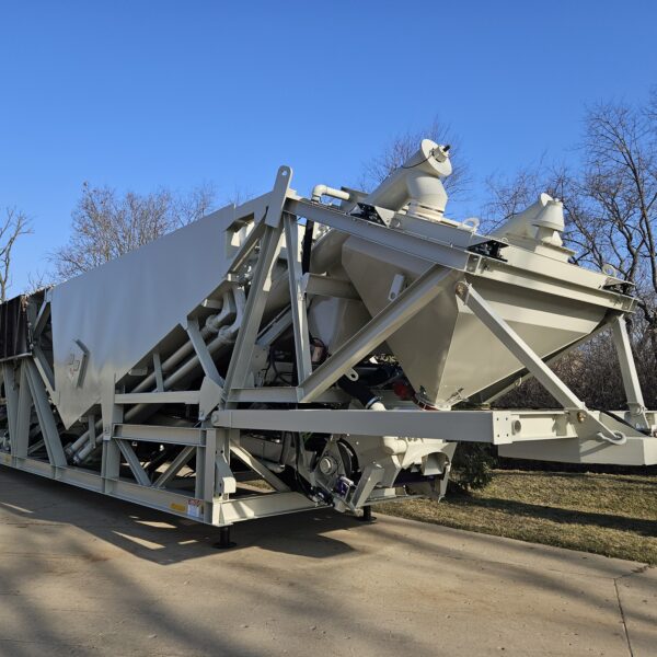 Large industrial concrete batching plant with multiple aggregate compartments and and large batch belt parked on a paved surface with trees and blue sky in the background.