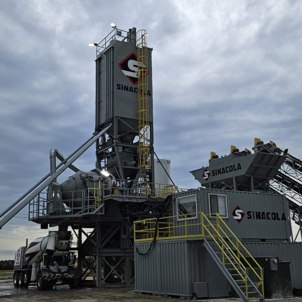 A large concrete batching plant with “Sinacola” logos on tall silos and equipment, surrounded by metal stairs and railings, stands under a cloudy sky on a gravel lot. A cement mixer truck is parked nearby.