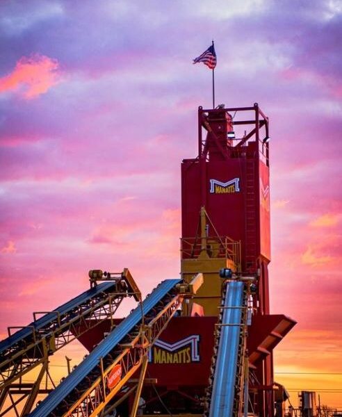 A red industrial structure labeled Manatts stands against a vibrant sunset sky, with an American flag at the top and conveyor belts extending into the foreground.