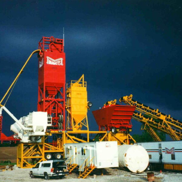 A brightly colored industrial plant with red, yellow, and white equipment stands under a dramatic dark sky. Vehicles, trailers, and a white truck are parked on the gravel in front.