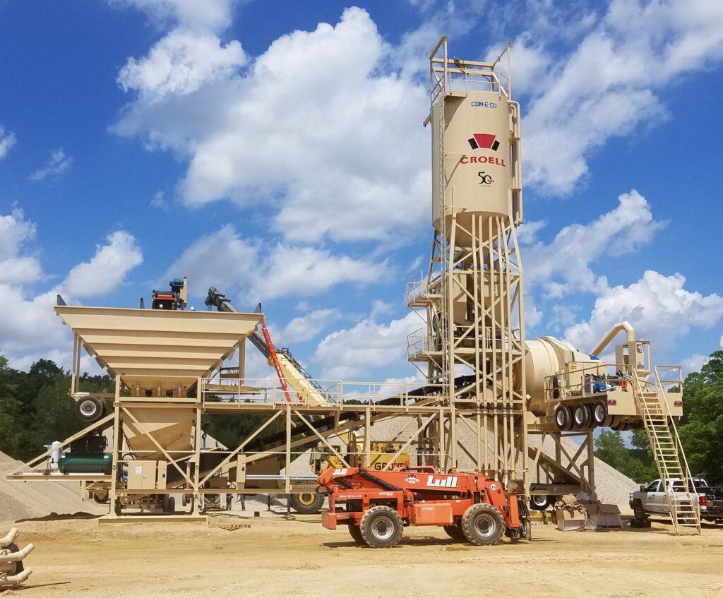 A concrete batching plant with a tall tan silo labeled Croell and various conveyor belts and machinery. A red forklift is parked in front, and workers are visible on the platform beneath a blue sky with white clouds.