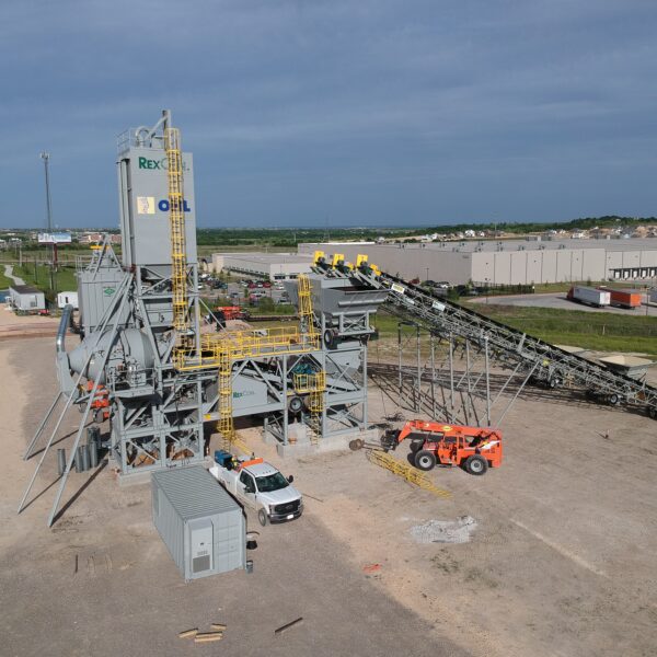 A large industrial concrete plant with conveyor belts and machinery stands on a dirt lot. Nearby are white and orange vehicles, with a warehouse and trucks visible in the background under a partly cloudy sky.