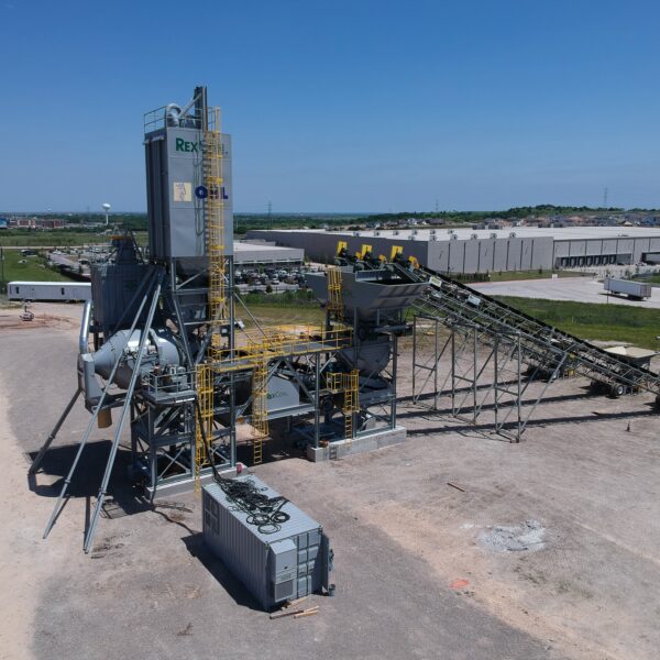 A large industrial asphalt plant with conveyor belts and storage units stands on a construction site, surrounded by dirt, roads, and nearby warehouses under a clear blue sky.