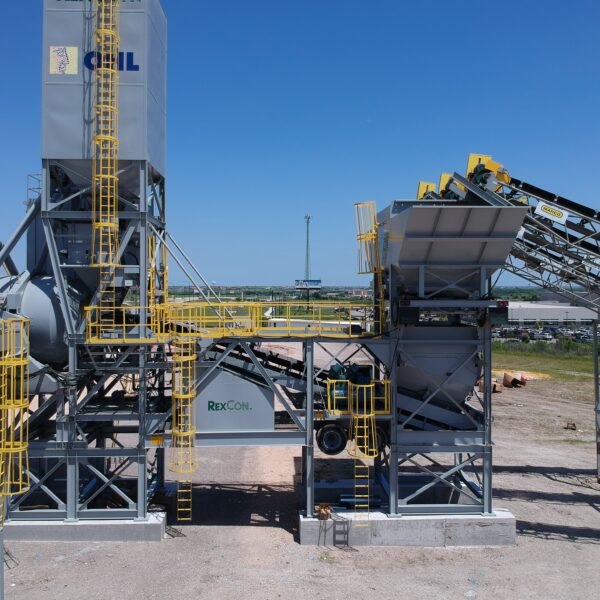 A large industrial concrete batching plant with silos, conveyor belts, and metal structures stands outdoors under a clear blue sky on a gravel lot. The equipment is painted gray and yellow.