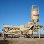 A large beige concrete batching plant with conveyor belts and a cylindrical silo stands on a construction site under a clear blue sky. The equipment is labeled P.W. Feenstra Construction.