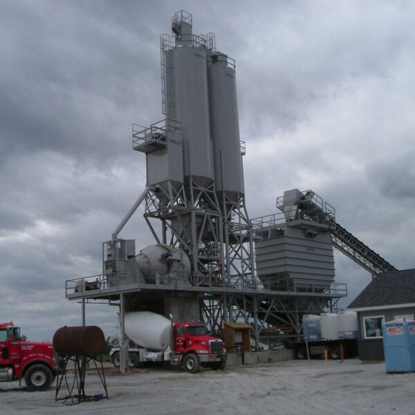 A concrete batching plant with tall silos, conveyor belt, and several red cement trucks parked nearby, under a cloudy sky. A small building and portable toilets are also visible on the gravel lot.