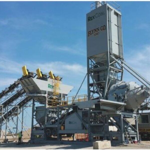 Large asphalt paving plant with conveyors, silos, and machinery stationed outdoors on a construction site under a clear sky. Equipment is labeled REXCON and FLYNN CO.