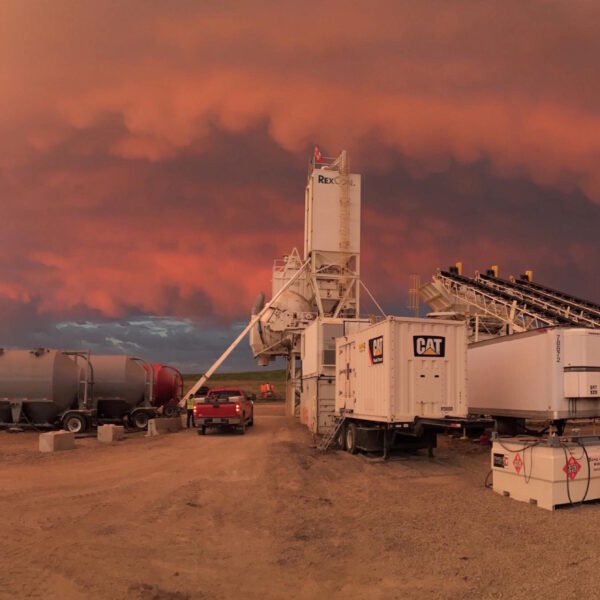 A construction site at sunset with large machinery, trailers, and a conveyor belt under a dramatic sky filled with dark, swirling orange and purple clouds.