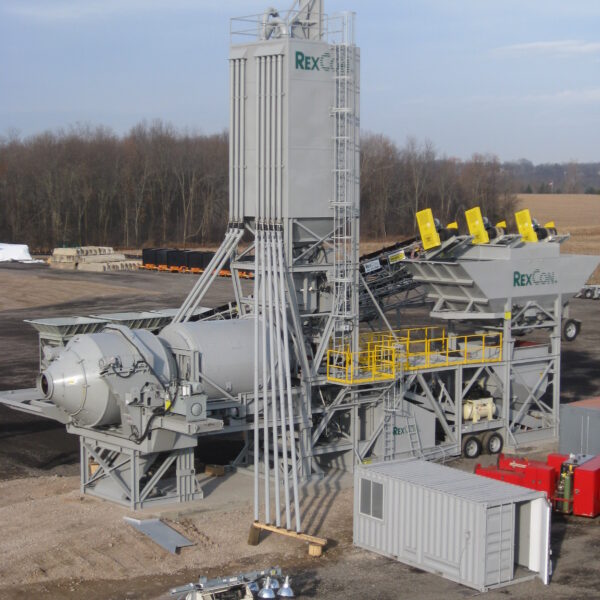A large gray concrete batching plant with multiple silos, conveyors, and machinery stands on a construction site with dirt ground and trees in the background. A white utility vehicle and storage containers are nearby.