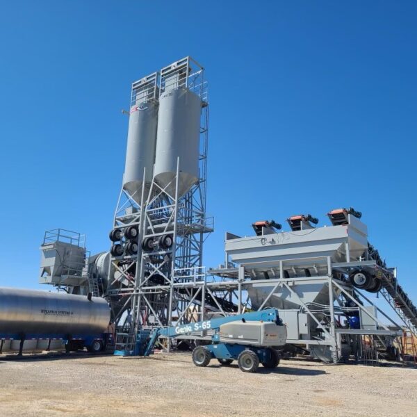 A large industrial concrete mixing plant with tall silos, conveyors, machinery, and a blue sky background, situated on a gravel lot with a tanker and construction equipment nearby.