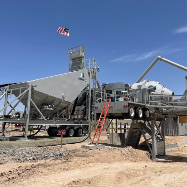 A large, mobile concrete mixing plant with an American flag flying on top stands on a construction site under a clear blue sky. Conveyor belts and ladders are visible, with dirt and machinery in the foreground.