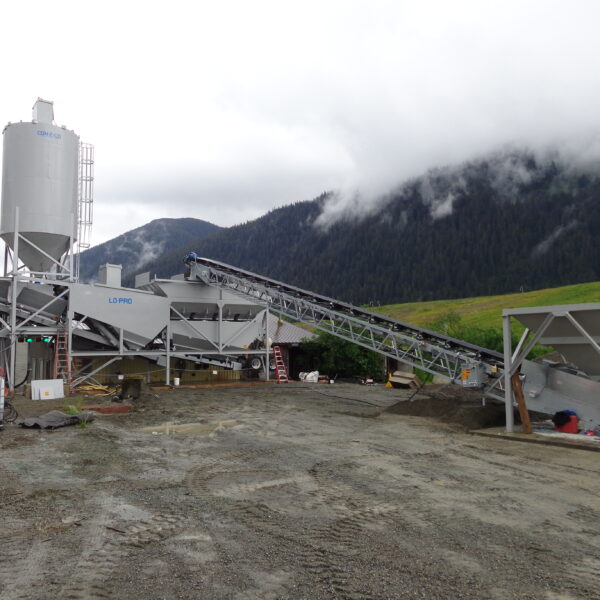A concrete batching plant with a large silo, conveyor belt, and equipment sits on a gravel lot. Misty mountains and grassy hills are visible in the background under a cloudy sky.