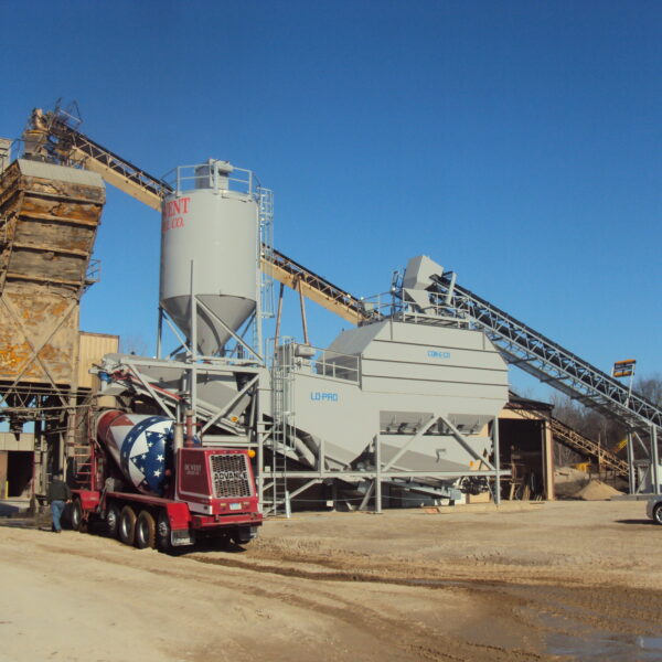 A concrete batching plant with silos, conveyor belts, and a red cement truck featuring a star and stripes design parked in front, under a clear blue sky. Sand and gravel piles are visible in the background.