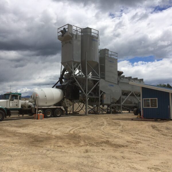 A concrete batching plant with silos, conveyors, and mixing trucks parked nearby. A small blue office building sits to the right, all on a dirt lot under a cloudy sky.