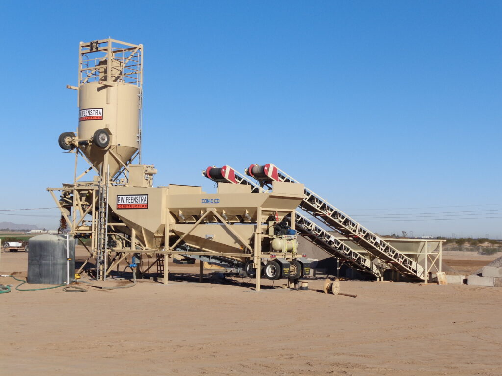 A beige concrete batching plant with conveyors and silos stands on a sandy construction site under a clear blue sky. Equipment and tanks are visible nearby.