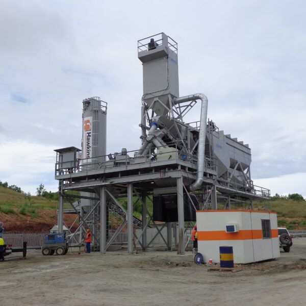 A large asphalt mixing plant with silos and machinery on a construction site. Workers in safety gear and equipment, including a generator and vehicles, are visible around the structure. Trees and cloudy sky in the background.