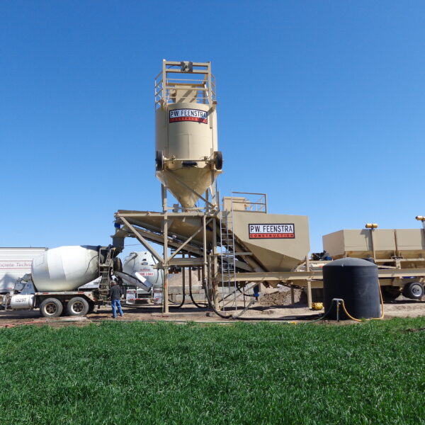 A concrete batching plant with large silos labeled PW FEENSTRA, a cement mixer truck, and workers on a construction site under a clear blue sky, with green grass in the foreground.