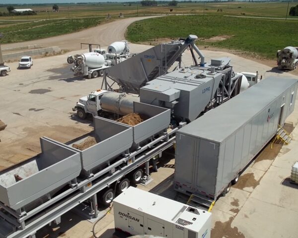 A large mobile concrete batching plant with multiple hoppers for materials is set up on a paved lot. Trucks, a trailer, and equipment surround the plant, with fields and roads visible in the background.