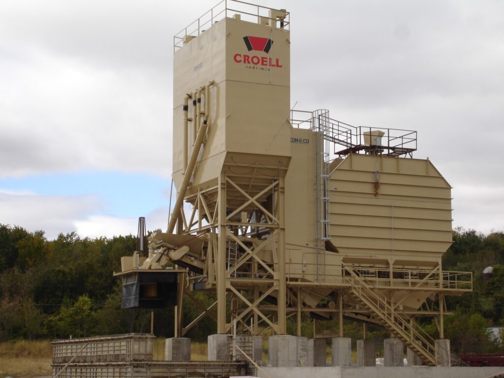 A large beige industrial concrete batching plant with the Croell logo stands outdoors on a concrete base, surrounded by greenery under a cloudy sky.
