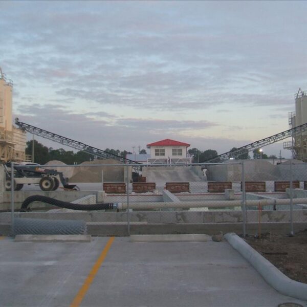 A construction site with two tall silos connected by conveyor belts, a fenced-off area with building materials, and a red-roofed building in the background under a cloudy sky.