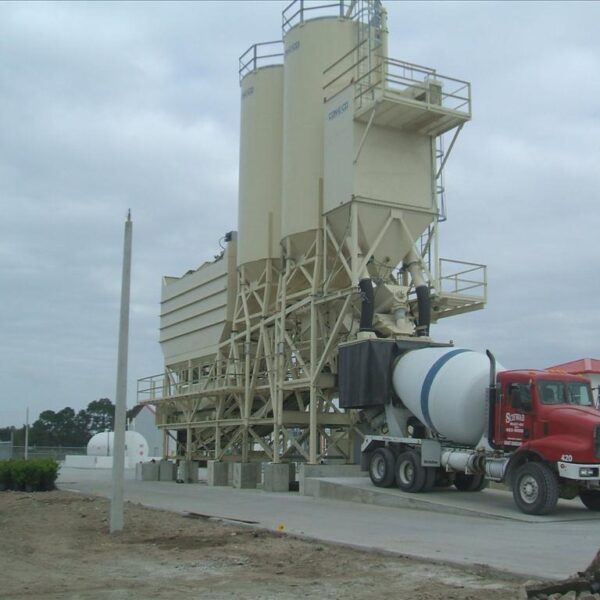 A cement mixer truck is parked next to a large beige concrete batching plant on a paved site, with cloudy skies overhead and some trees and a building in the background.