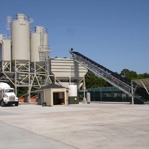 A concrete batching plant with large silos, conveyor belt, and a cement mixer truck on a paved lot, surrounded by small buildings and greenery under a clear blue sky.