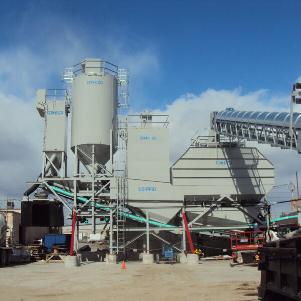 A concrete batching plant with tall silos, conveyor belts, and industrial equipment is set up outdoors under a partly cloudy sky. A cement truck is parked nearby on the dusty ground.