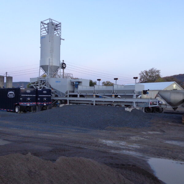 A concrete batching plant with tall silos, storage bins, and conveyor belts stands on a gravel lot. A black truck and a yellow bulldozer are parked nearby under a clear sky with trees and buildings in the background.
