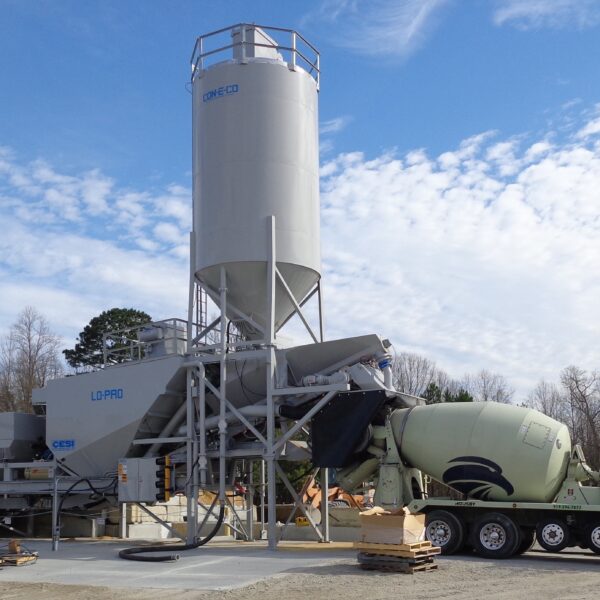 A concrete batching plant with large silos and conveyor systems operates outdoors as a cement mixer truck collects ready-mix concrete. Trees and blue sky with scattered clouds are in the background.